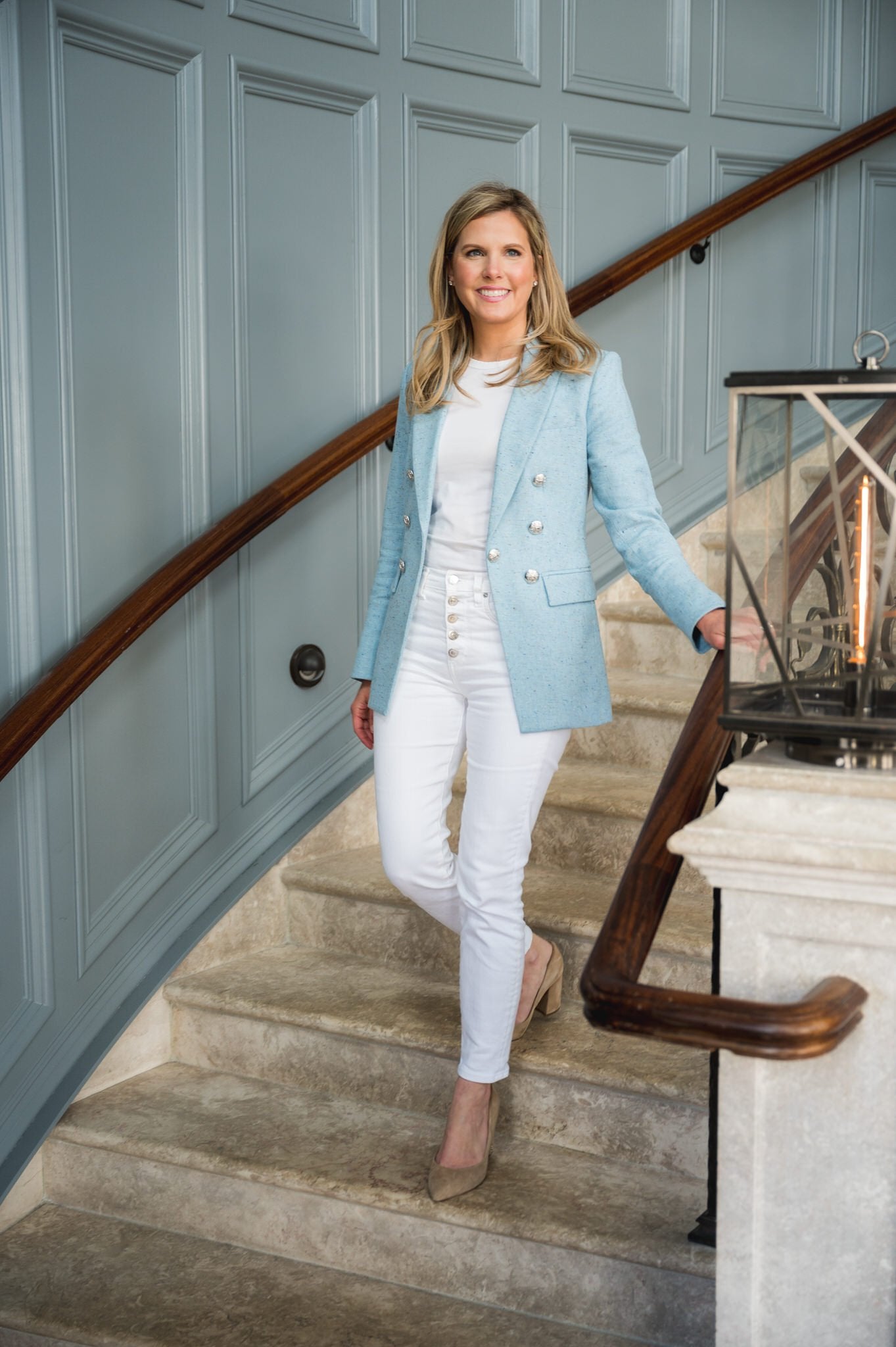 Woman in a light blue blazer and white pants standing on a staircase with wooden railings.